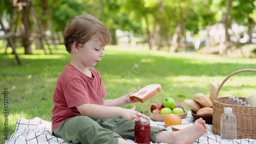 Adorable Caucasian boy is sitting on the grass at the village park, hungry, dipping his fingers into a can of jam. Then I spread bread with the intention of eating it. family vacation Concept.