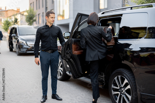 Male driver helps a business lady to get in a car, opening door of a luxury SUV taxi. Business lady with handbag wearing black formal wear. Concept of transportation service © rh2010
