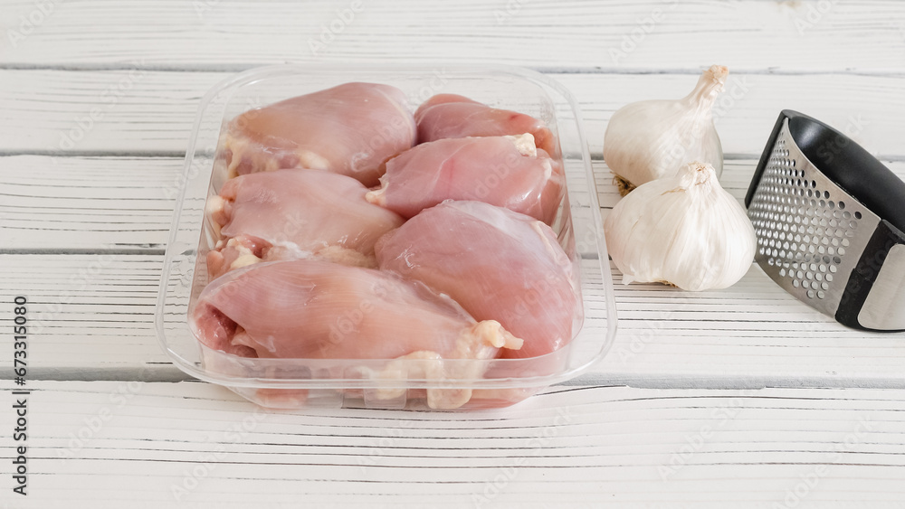 Raw chicken thighs in a plastic container, and fresh garlic and garlic press close-up on a white wooden board with copy space