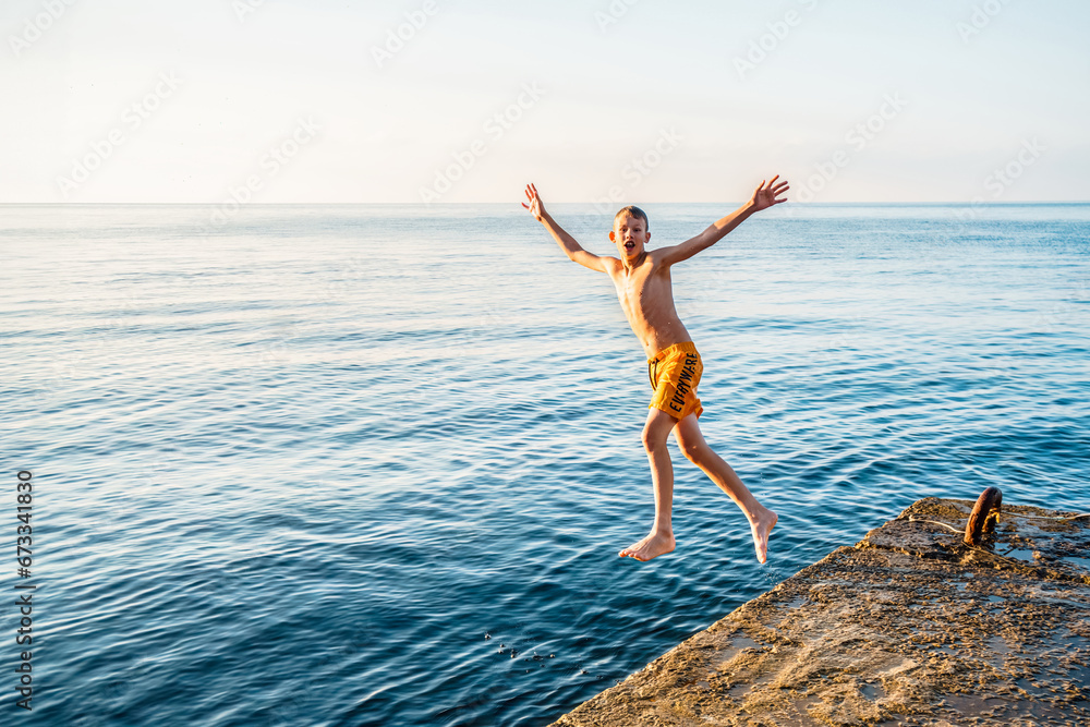 Healthy boy jumps into tranquil ocean water from stone pier in sunny morning. Sportive child dives in sea at summer resort. Active lifestyle
