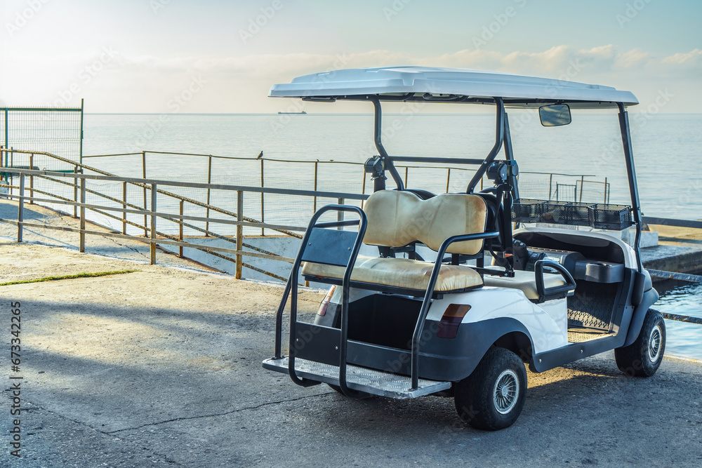 Comfortable golf cart parked on city waterfront by tranquil sea. Small tourist car on stone embankment near ocean bay. Compact vehicle at resort
