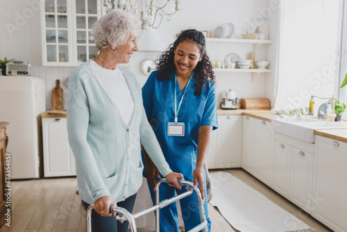 African american medical nurse volunteer helping senior female at home, teaching her to walk with walker, smiling and giving instructions how to use device, standing together in cozy kitchen