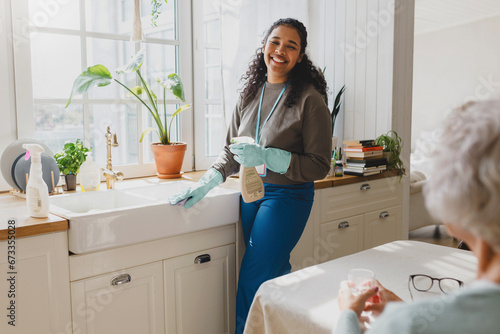 Selective focus on happy african american volunteer helping senior lady to clean house, standing in kitchen in raisin gloves washing sink with detergent, talking to elderly female sitting at table
