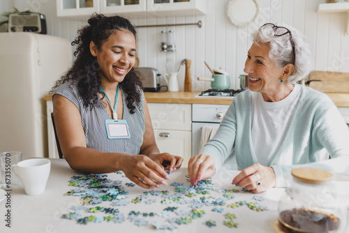 Cheerful senior lady having fun plying puzzle game at kitchen table with black female volunteer, taking care of her as social support of retired persons, spending leisure time, talking photo