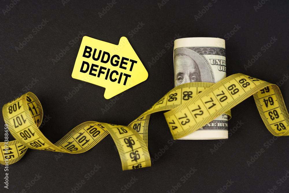 On a black surface there are dollars wrapped in measuring tape and stickers with the inscription - budget deficit - obrazy, fototapety, plakaty 