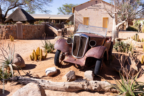 Old car wreck in front of the modest 1951 Immanuel Church in the remote town of Solitaire, Namibia, seen during a very bright sunny morning photo