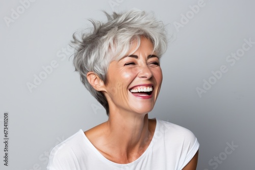Vibrant mature woman with silver hair laughing joyously on a grey background, white smile.
