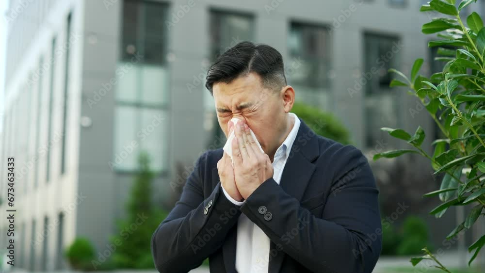 Sick asian businessman in formal suit suffering from seasonal allergies on the street near an office building. Allergic male entrepreneur sneezes and wipes his nose with a handkerchief. Close up