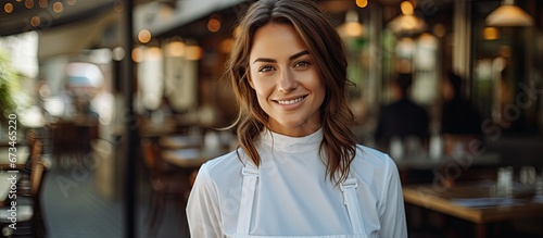 A smiling woman wearing a white blouse happily crosses her arms while standing on a blurred background of a restaurant terrace during her break from remote work