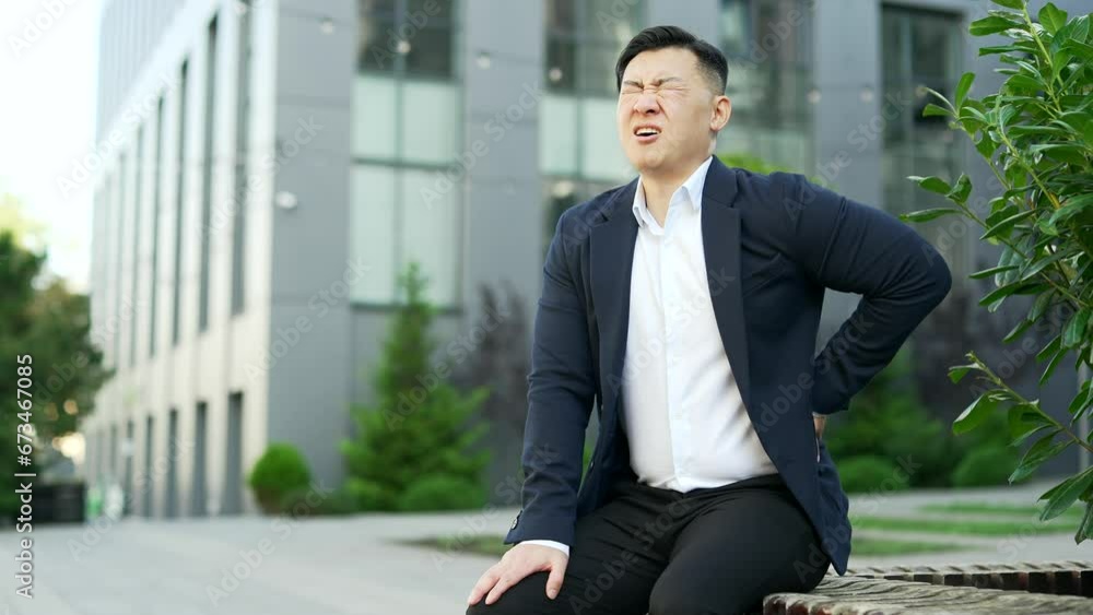 Tired overworked asian businessman in formal suit suffering from back pain sitting on a bench on the street near an office building. Upset exhausted entrepreneur massages painful lower back muscles
