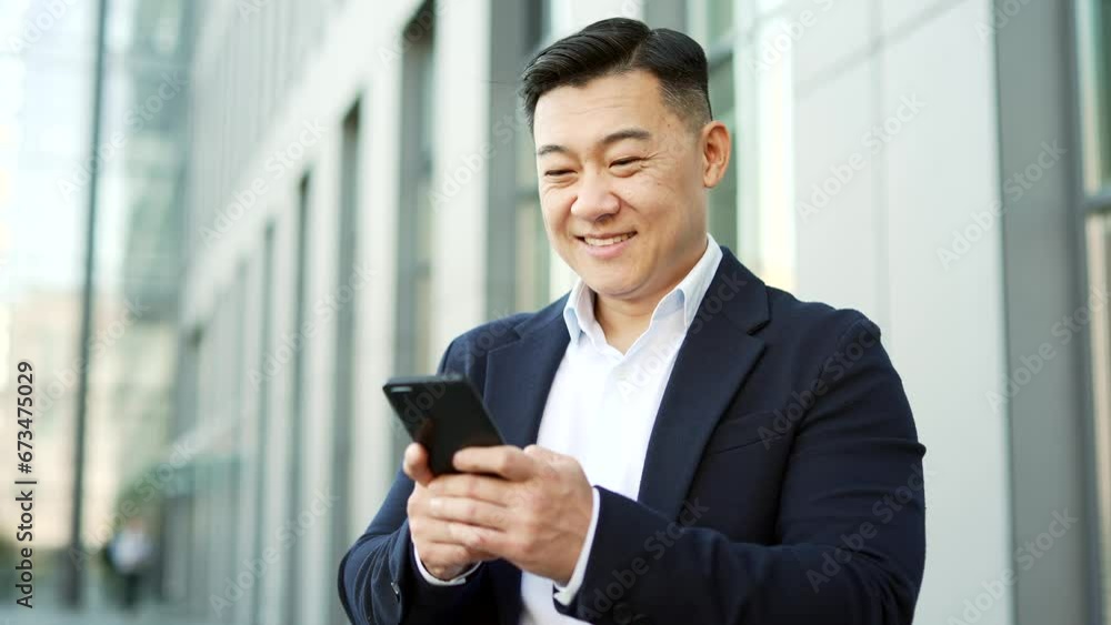 Smiling businessman in formal suit is using a smartphone while standing on street near an office building. Happy entrepreneur reads writes an email message or chatting online with a business partner Smiling businessman in formal suit is using a smartphone while standing on street near an office building. Happy entrepreneur reads writes an email message or chatting online with a business partner