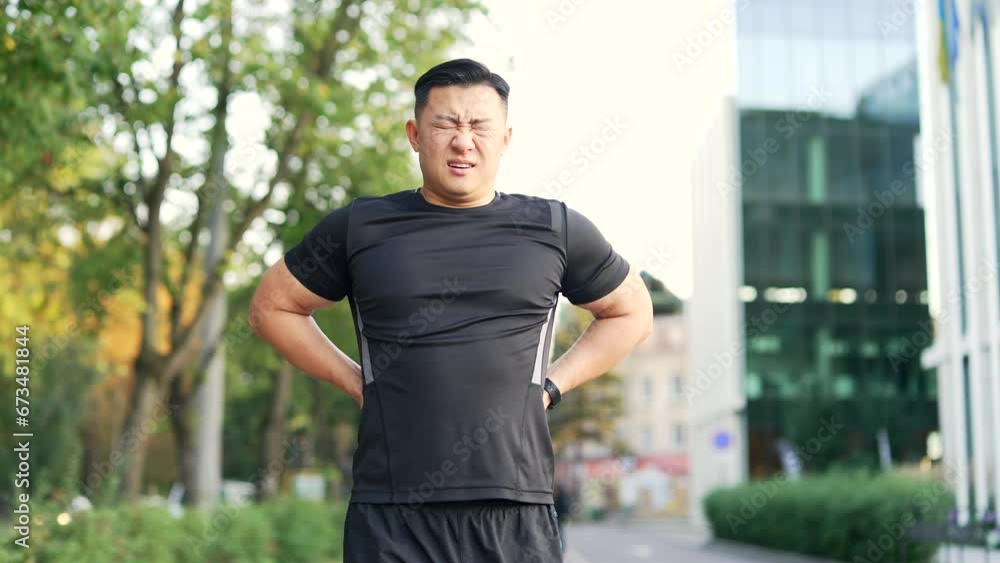 Adult asian runner suffering from back pain, sports injury, muscle spasm in city street. Man in sportswear having back flank ache and problem after exercise jogging outside outdoors in urban city park Adult asian runner suffering from back pain, sports injury, muscle spasm in city street. Man in sportswear having back flank ache and problem after exercise jogging outside outdoors in urban city park