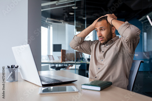 Frustrated and sad man at workplace reading online message from laptop, businessman sitting inside office at workplace, desperate unhappy, got bad news, bankruptcy, layoff.