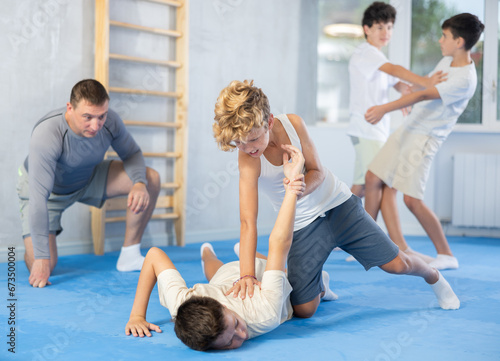 Teenage students taking self-defense classes at training center. Boy applying armlock technique to face-down opponent under careful supervision of instructor..