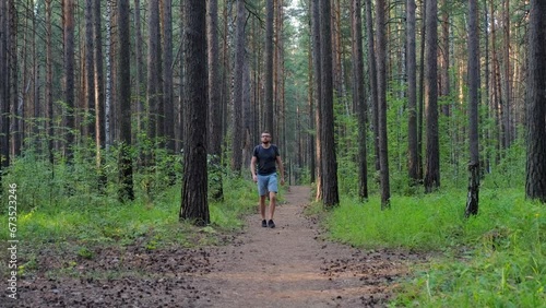 man in glasses, blue shorts and a gray T-shirt with a backpack walks through a pine forest, having rest outdoors at sunset.