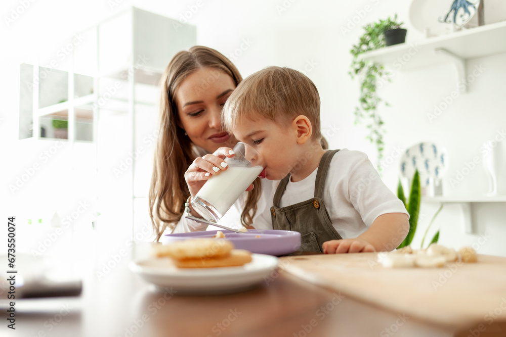 mother feeds her little son at home in the kitchen, woman gives glass of milk for breakfast, hungry boy drinks milk mother feeds her little son at home in the kitchen, woman gives glass of milk for breakfast, hungry boy drinks milk