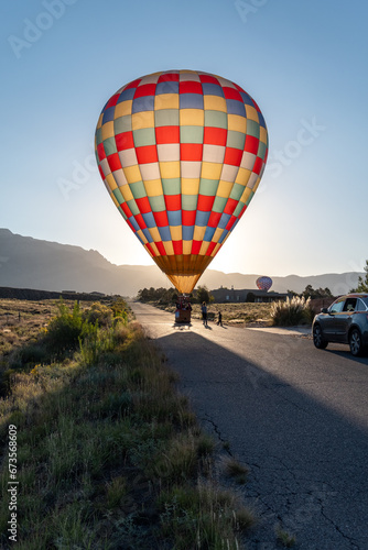 A colorful hot air balloon lands on a road in Albuquerque, New Mexico during the Balloon Fiesta. The balloon is lit like stained glass by the morning sun. The road leads toward the mountains behind.