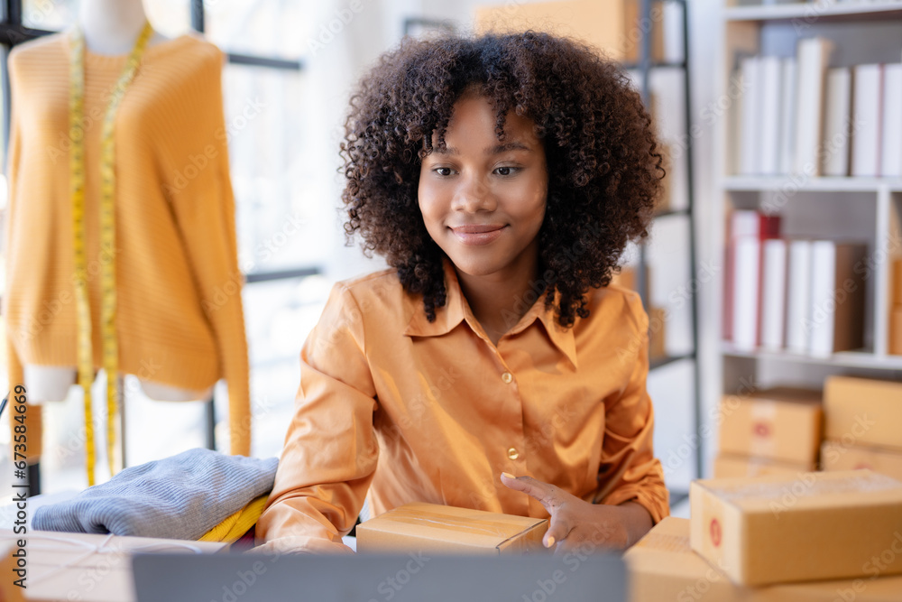 Fototapeta premium Young African American woman online selling. Startup small business entrepreneur or freelance african woman using a laptop with box. Online marketing packaging box and delivery, SME concept.