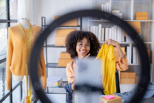 Young African American vlogger woman influencer. Smiling retail store owner enjoys selling live items online. Woman using mobile phone video call for sell clothes live stream selling online. photo