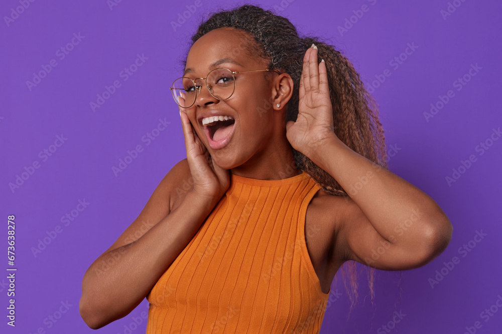 Young cheerful excited African American woman is surprised and puts palm to ear to hear answer to question or new shocking news dressed in casual clothes stands on plain purple background. - obrazy, fototapety, plakaty 