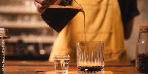 A female barista is pouring an americano or filter coffee to a glass. Concept of vintage cafe or coffee shop. Close up of hand pour black coffee ready to serve. Preparation, aroma and smoke. photo