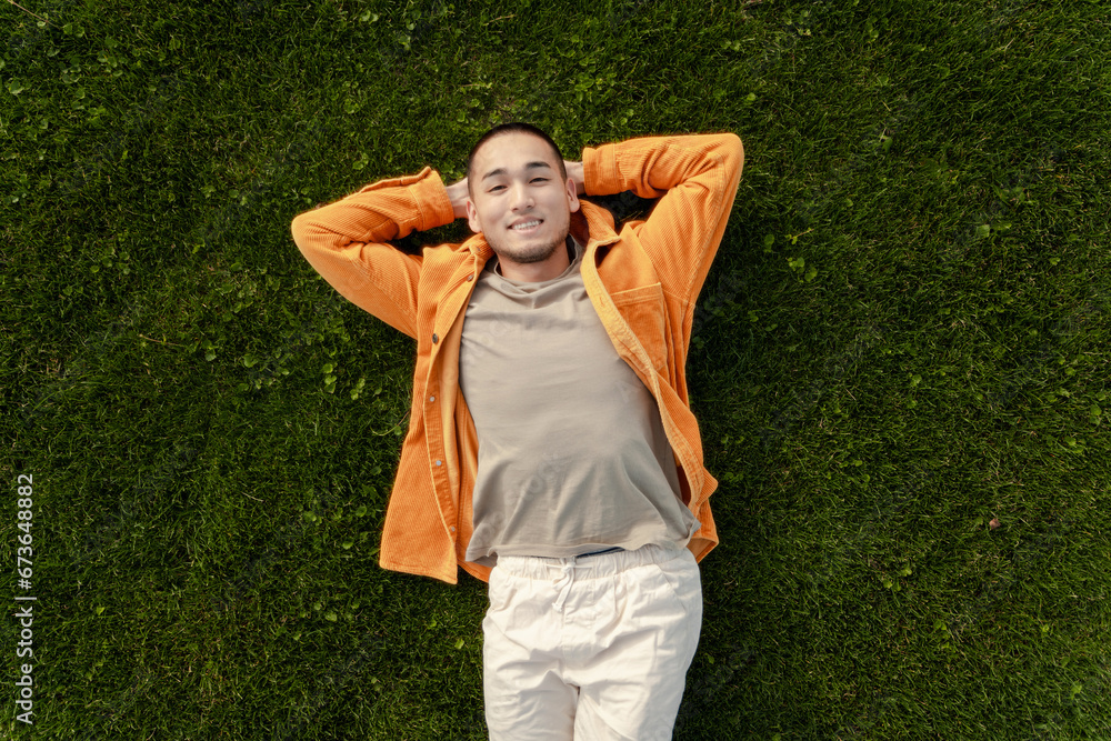 Positive Asian man in stylish casual clothes lying on green grass, relaxing looking at camera Positive Asian man in stylish casual clothes lying on green grass, relaxing looking at camera