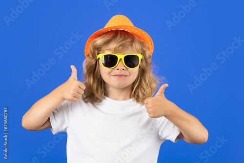 Kid boy making thumbs up sign. Fashion portrait of kid in summer hat, t-shirt and sunglasses on blue studio isolated background.