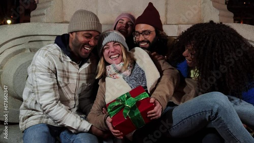 Excited young adult woman holding in her arms a birthday gift or christmas present of her best friends laughing with complicity together. Happy group of multiracial people celebrating the xmas night photo
