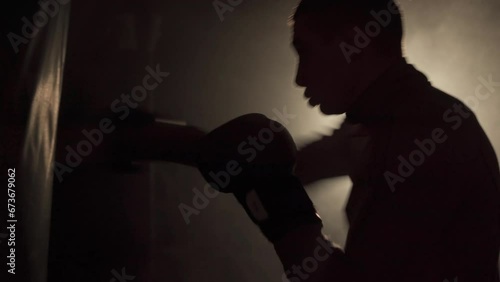 Male boxer training with punching bag in dark sports hall. Exercise for the big fight. Boxer hits