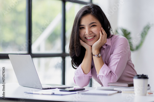 Young Asian woman smiling reads the screen of a laptop computer while relaxing working on a comfortable place by the wooden table at office. Excited happy Asian woman.