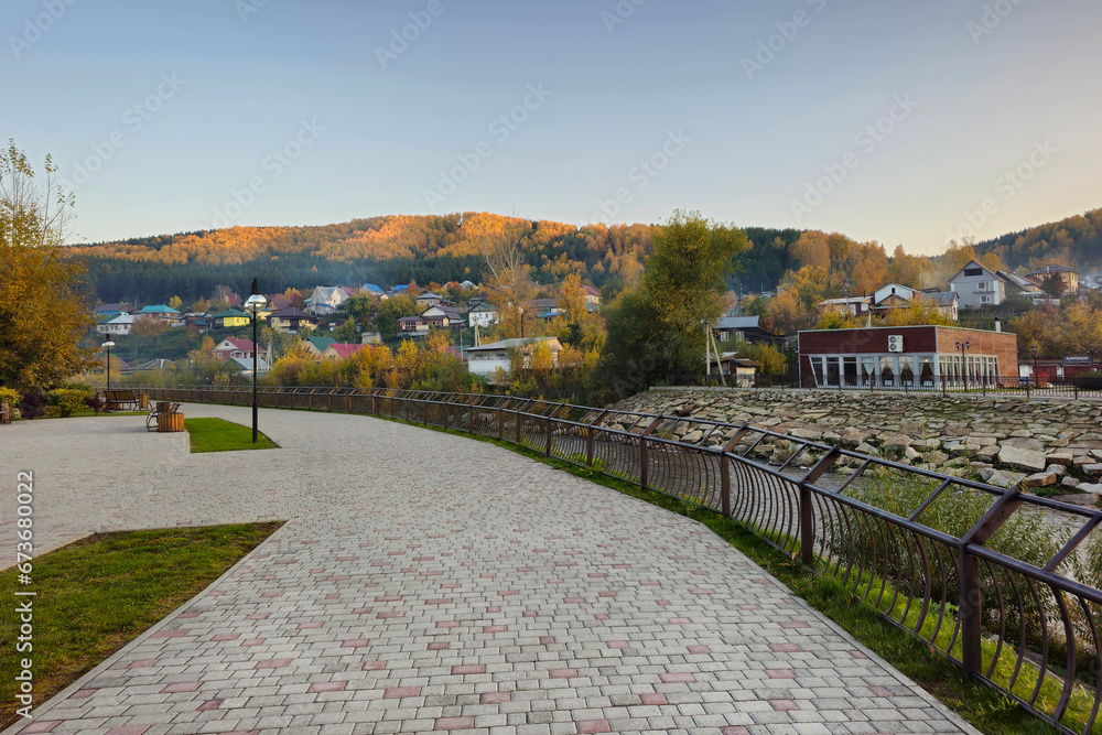 Tile embankment with railings along the river. Evening village on the banks of a mountain river. Rustic landscape.  Rocky bed of the river flowing through the village. Gorno-Altaisk.
