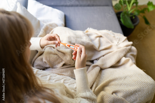 Woman checking digital thermometer sitting on sofa at home photo