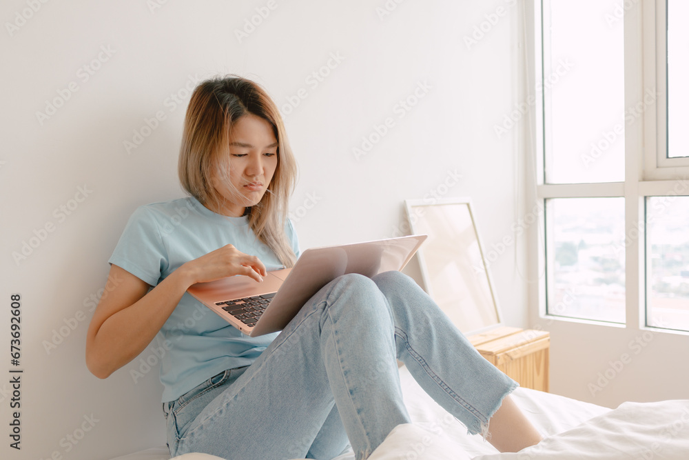 Unhappy asian Thai woman dislike and bored face, using laptop for working at home, putting computer on pillow lap, sitting on white bed, worried trouble at apartment room alone. Unhappy asian Thai woman dislike and bored face, using laptop for working at home, putting computer on pillow lap, sitting on white bed, worried trouble at apartment room alone.