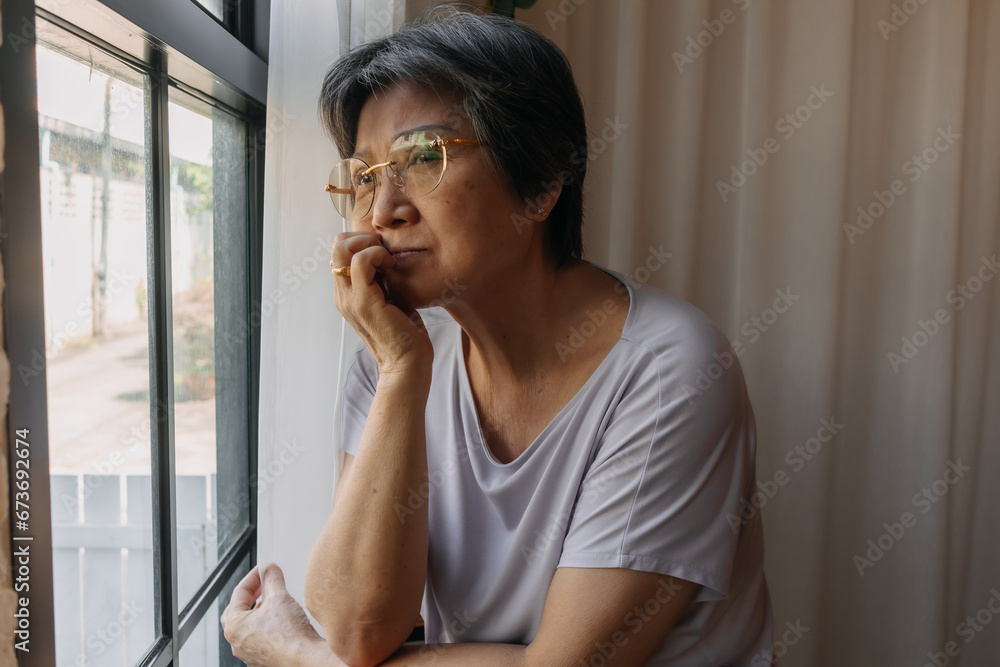 Asian old woman mother sitting on chair and looking out the window, resting chin on arm, missing and waiting for someone and thinking something sad, living alone. Asian old woman mother sitting on chair and looking out the window, resting chin on arm, missing and waiting for someone and thinking something sad, living alone.