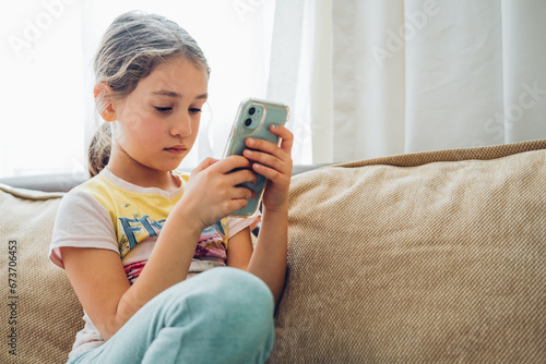 Cute little kid girl using digital tablet technology device lying on sofa alone. Small child hold computer surfing Internet play game at home. Children tech addiction concept. 