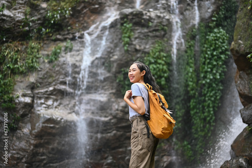 Image of smiling female tourist with backpack enjoying the view of beautiful tropical waterfall