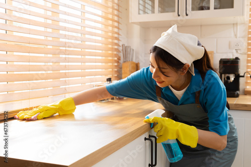 Cheerful Asian cleaning services worker female in uniform wearing apron and gloves standing in kitchen wiping table surface using hygiene sprayer, small business cleaning services company hiring woman