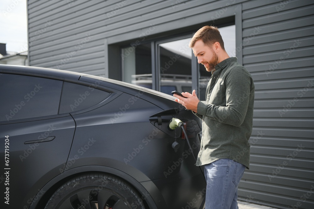 handsome bearded man in casual wear, standing at the charging station and charger for an electric car. Eco electric car concept handsome bearded man in casual wear, standing at the charging station and charger for an electric car. Eco electric car concept