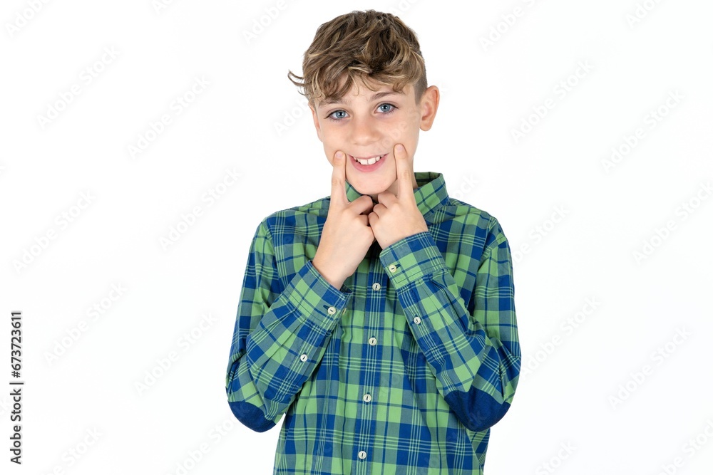 Happy caucasian kid boy wearing plaid shirt with toothy smile, keeps index fingers near mouth, fingers pointing and forcing cheerful smile Happy caucasian kid boy wearing plaid shirt with toothy smile, keeps index fingers near mouth, fingers pointing and forcing cheerful smile