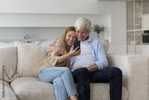 Happy mature dad hugging adult pretty daughter child with smartphone on couch, watching online media content, talking on video call, using online application for Internet communication