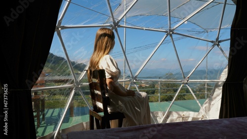 Woman seated in transparent dome tent, overlooking majestic mountain scenery, unique luxury camping experience, serene getaway. Travel and leisure.