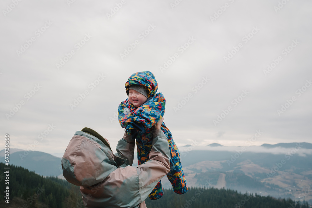 Mother throws up baby son in the sky on an autumn day. Family vacation with children in mountains. People stand and relax in country. Mom hugs child enjoy time together. Active weekend travel concept