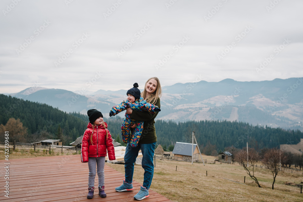 Mother throws up baby kid in the sky on a terrace on an autumn day. People stand in backyard of country house. Mom, daughter, hug son enjoy time together. Active weekend travel concept in mountains.