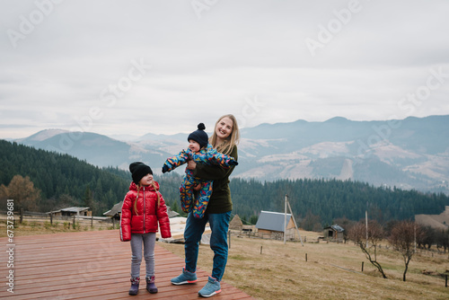 Mother throws up baby kid in the sky on a terrace on an autumn day. People stand in backyard of country house. Mom, daughter, hug son enjoy time together. Active weekend travel concept in mountains.