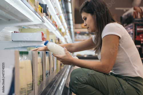 Young woman squatting in the daily fresh drinks section of the supermarket checking the nutritional properties of the milk bottle she is holding. Batch cooking concept.