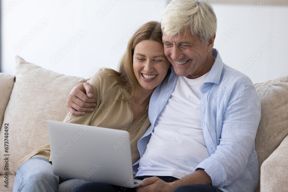 Happy senior father and adult daughter child watching movie on laptop at home, resting on couch with computer, hugging, laughing, looking at screen, enjoying family leisure, online communication