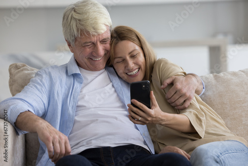 Happy beautiful adult daughter woman and senior father using online application, web service on smartphone, hugging on home sofa with love, affection, speaking on video call, taking selfie