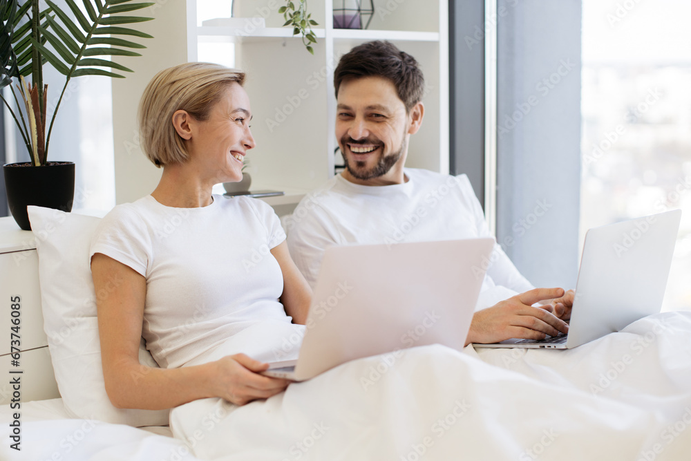 Cheerful amorous people accessing portable computers while spending time together in spacious apartment at weekend. Joyful caucasian family scrolling social media on wireless laptops. Cheerful amorous people accessing portable computers while spending time together in spacious apartment at weekend. Joyful caucasian family scrolling social media on wireless laptops.