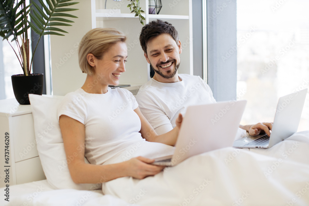 Beautiful young woman sitting in bed and sending text messages using modern laptop while her husband assisting her with advice. Loving married couple achieving tech-life balance resting at home.