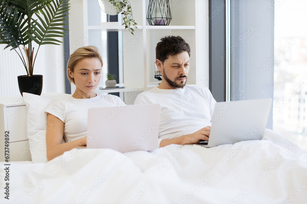 Happy caucasian married couple dealing with smart gadgets while relaxing under cozy duvet in airy bedroom of modern apartment. Cheerful family man and woman uploading content to social media. Happy caucasian married couple dealing with smart gadgets while relaxing under cozy duvet in airy bedroom of modern apartment. Cheerful family man and woman uploading content to social media.